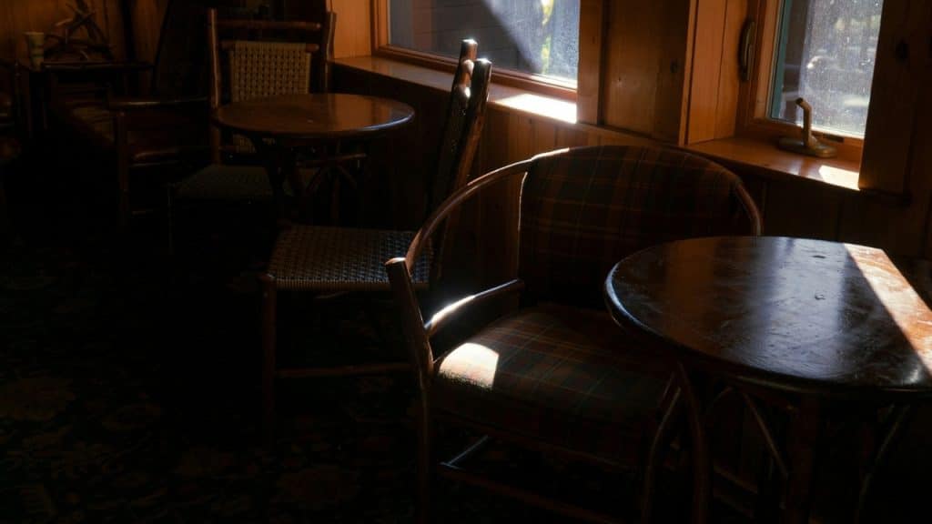 An empty wooden table and chairs sit in a dimly lit room near a window with sunlight.