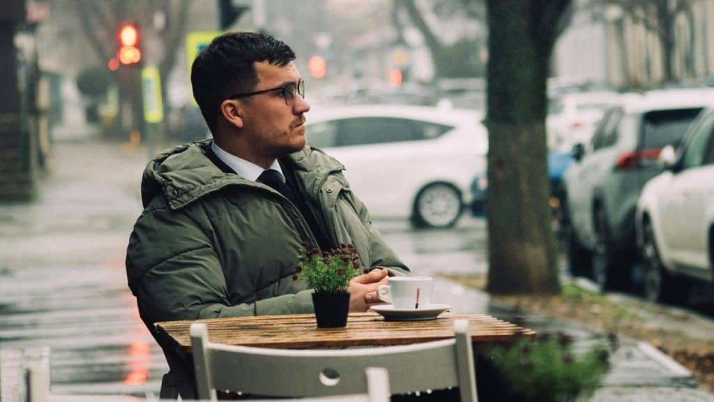 A man in a winter coat sits at an outdoor cafe table on a rainy day.