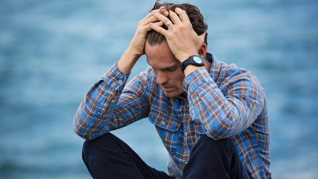 A man in a blue plaid shirt sits outside with both hands on his head.