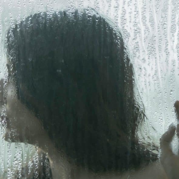 A woman standing behind a water-soaked glass and touching it with her hand as she looks to the side.
