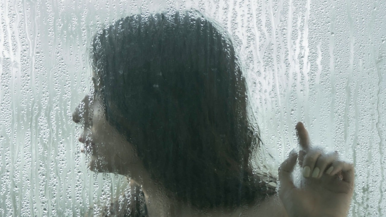 A woman standing behind a water-soaked glass and touching it with her hand as she looks to the side.
