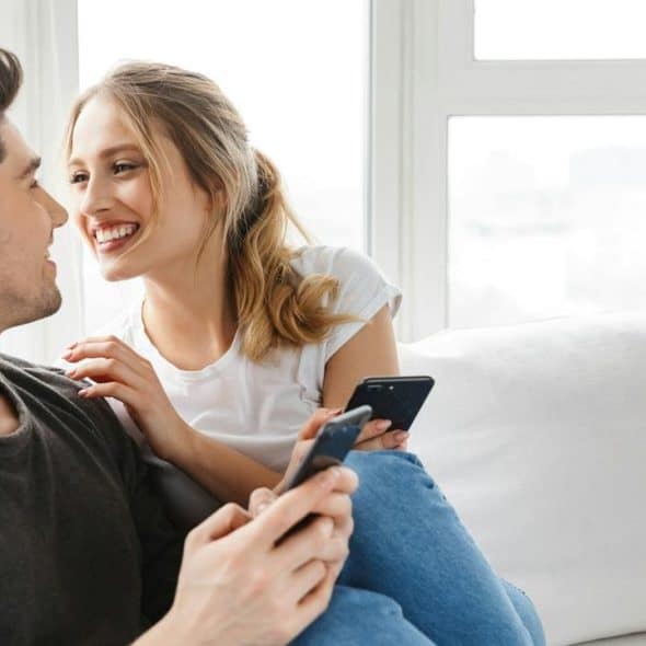 A smiling couple sitting on a couch with smartphones.