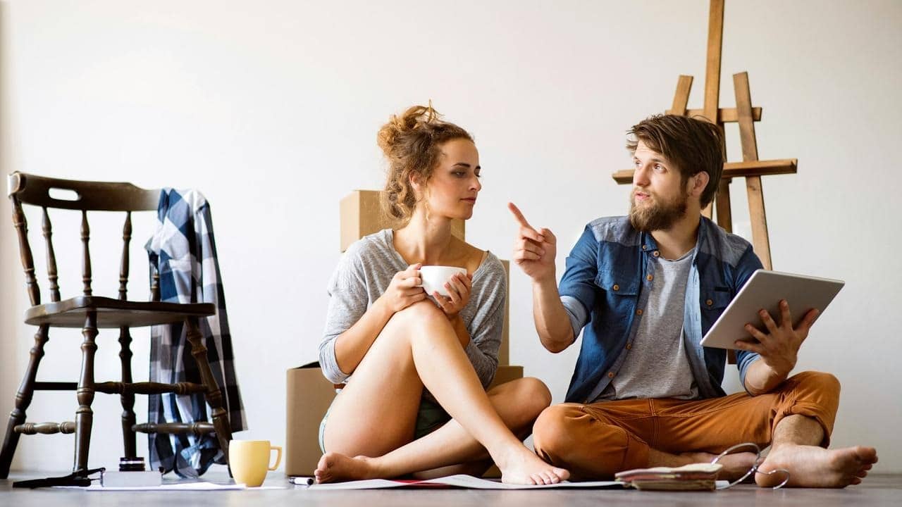 A couple sitting on the floor talking while holding a tablet and a cup.