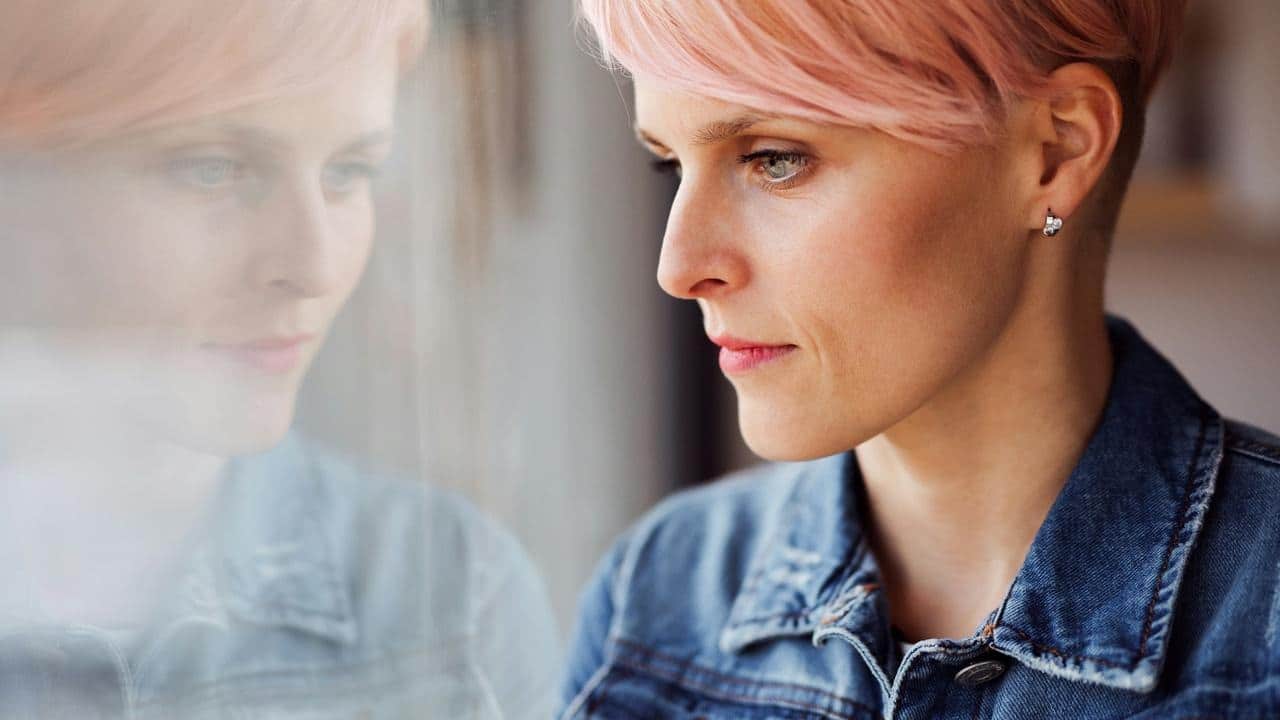 A woman with pink hair looking thoughtfully out a window.