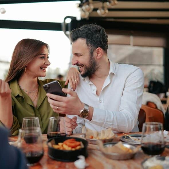 A smiling couple sitting at a table while the bearded man shows something to a woman on his phone who looks at him while holding her sunglasses with her free hand.