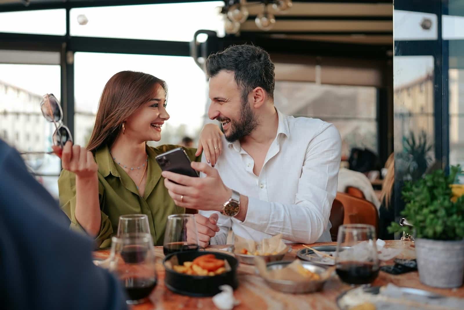 A smiling couple sitting at a table while the bearded man shows something to a woman on his phone who looks at him while holding her sunglasses with her free hand.