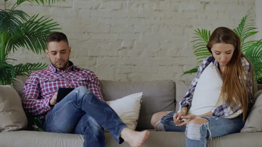 A couple is sitting separated on a sofa, both looking down and engrossed in their own activities