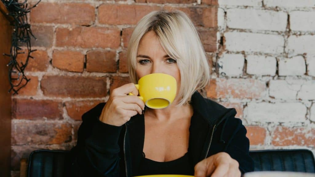 A blonde woman in a black jacket drinks from a bright yellow mug against brick.