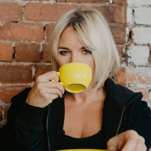 A blonde woman in a black jacket drinks from a bright yellow mug against brick.