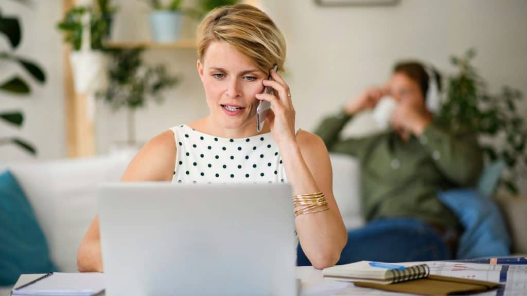 A woman talks on a mobile phone while using a laptop as a man sits blurred behind her.