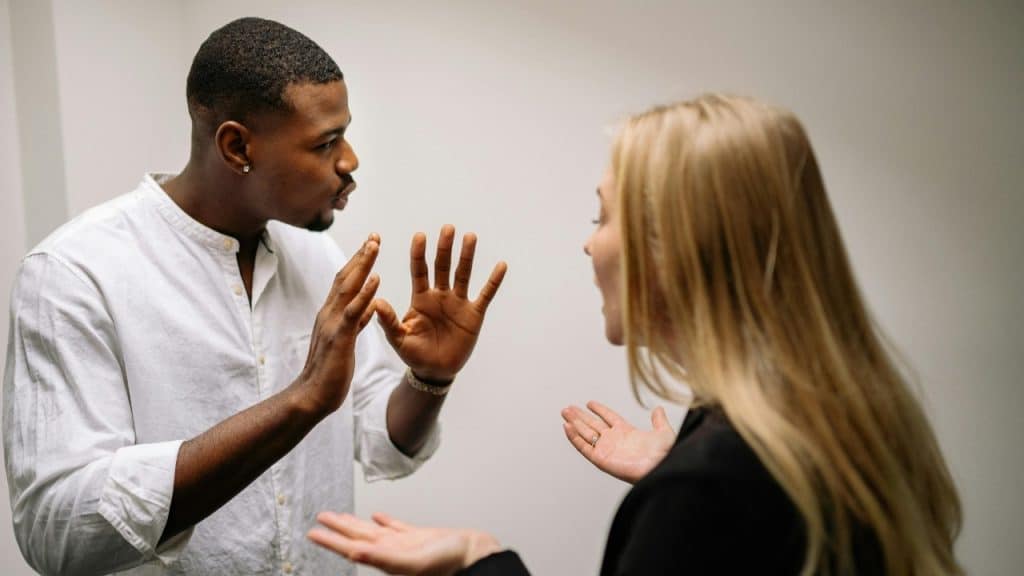A man and woman stand facing each other while gesturing with their hands during conversation.