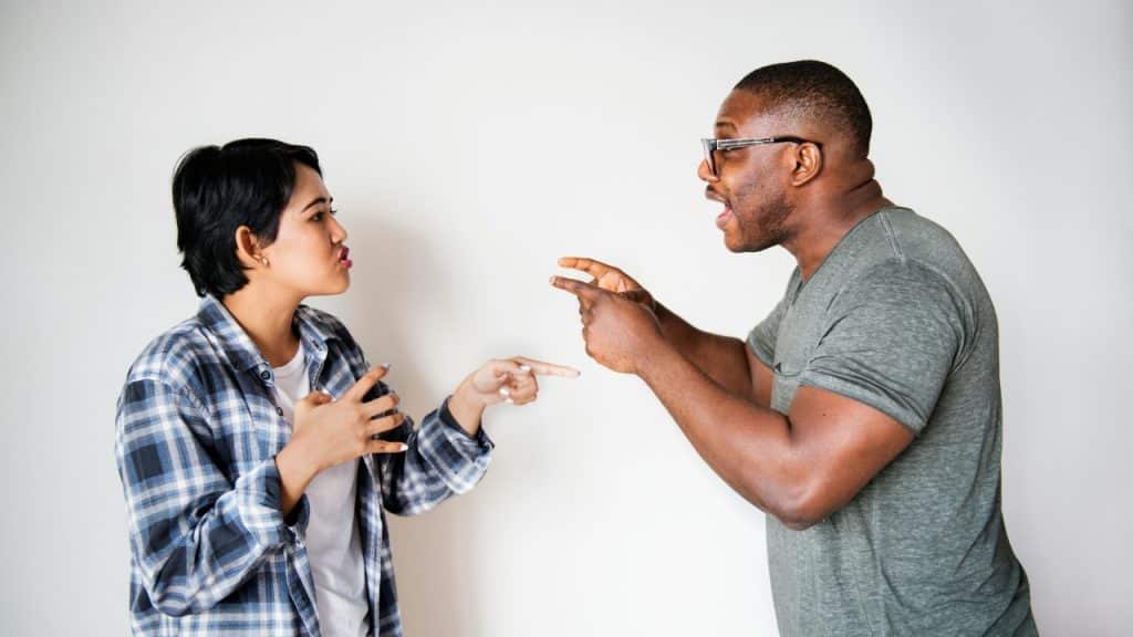 A man and woman point at each other while speaking in front of a white wall.