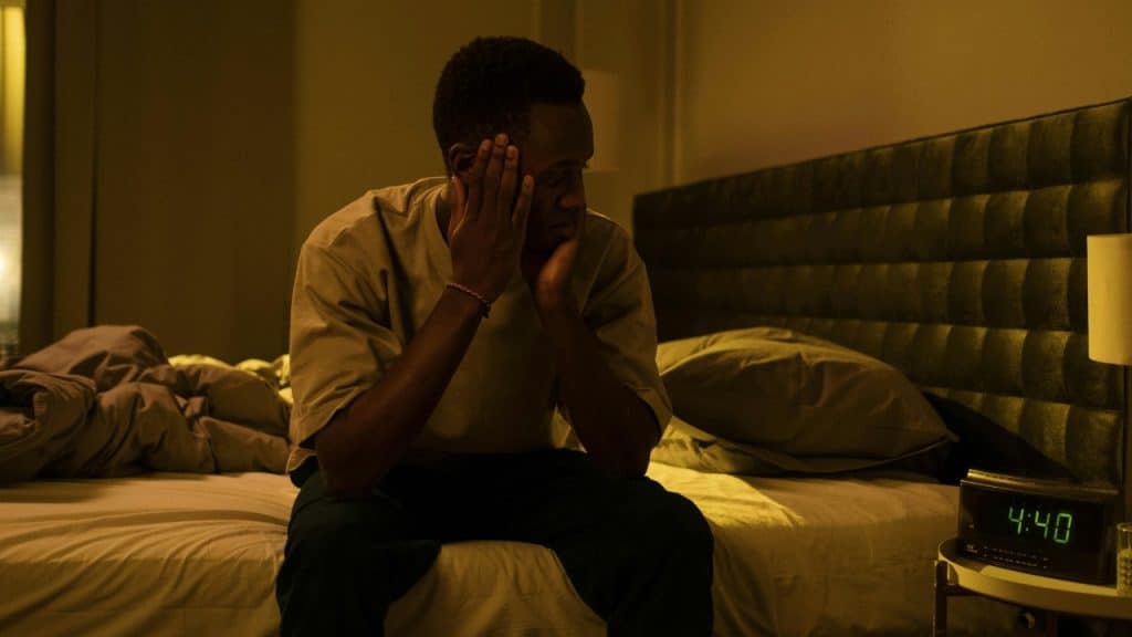 A man sits on a bed with his hands on his head near a digital clock.