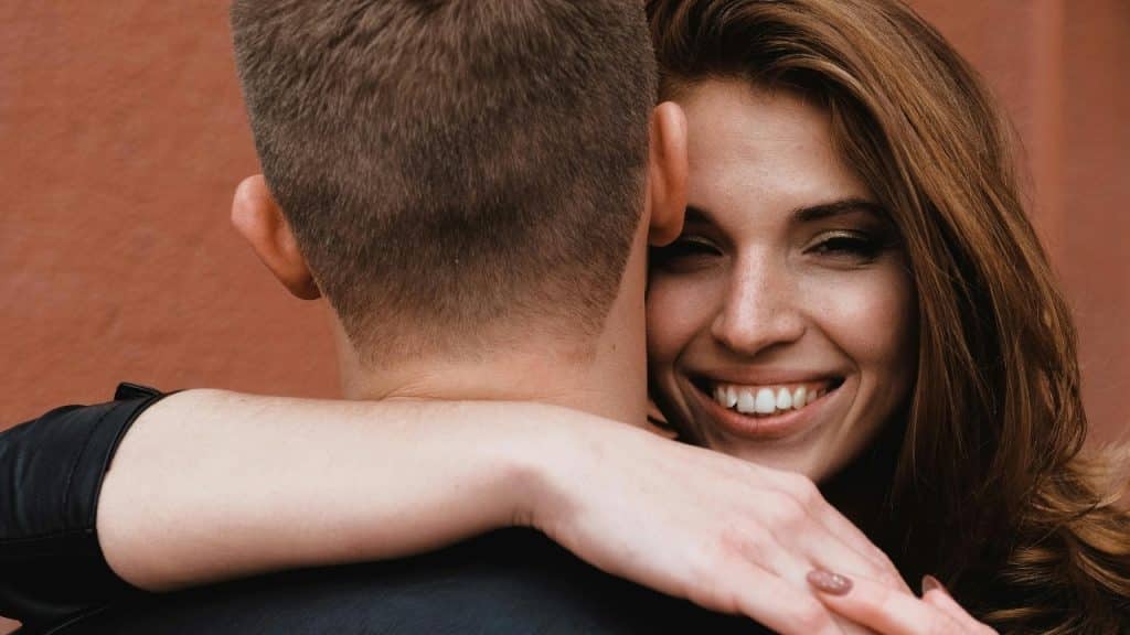 A woman with long brown hair smiles while hugging a man from behind his head.