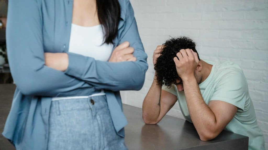 A person stands with arms crossed in front of a person sitting with head down.