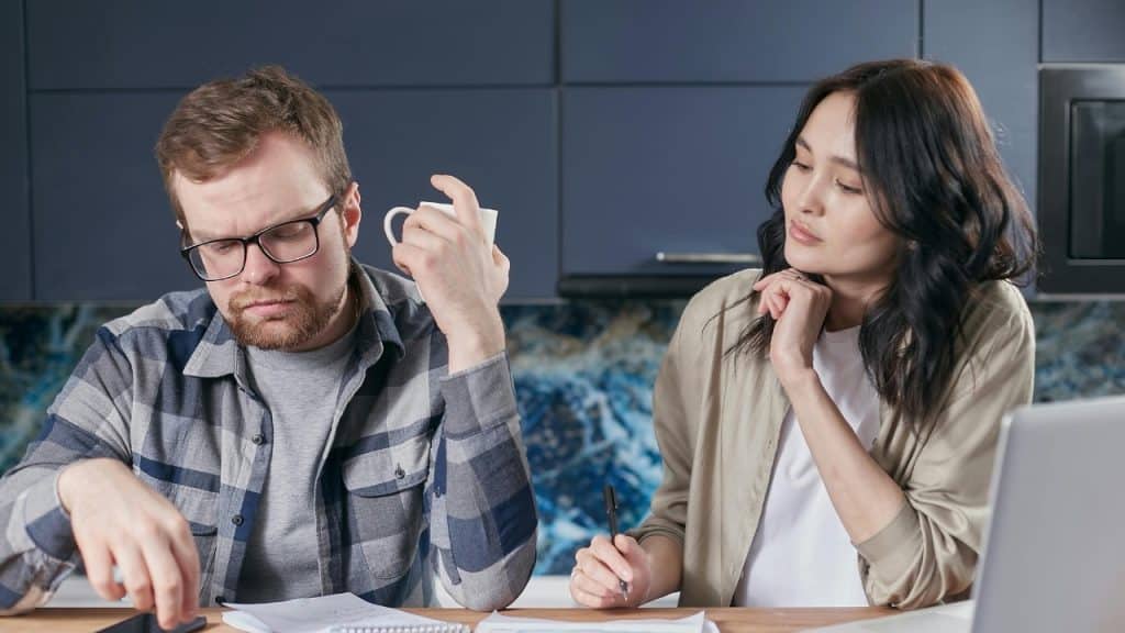 A man and woman sit at a table with papers, a laptop, and a mug.