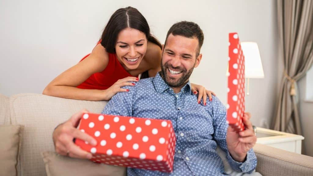 A smiling woman stands behind a man who is opening a red polka-dot gift box.