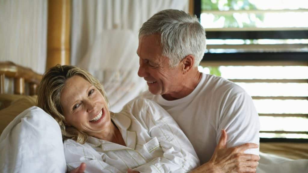 A smiling woman in white pajamas reclines in bed while an older man looks at her.