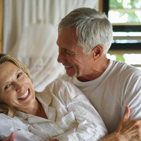 A smiling woman in white pajamas reclines in bed while an older man looks at her.