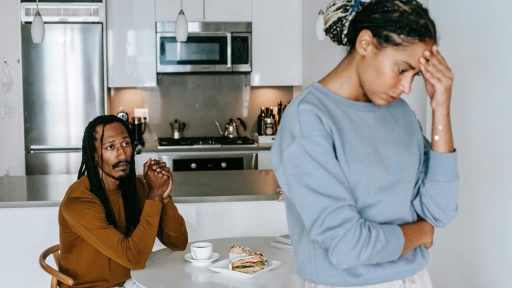 A man with dreadlocks sits at a table while a woman stands in front, holding her head.
