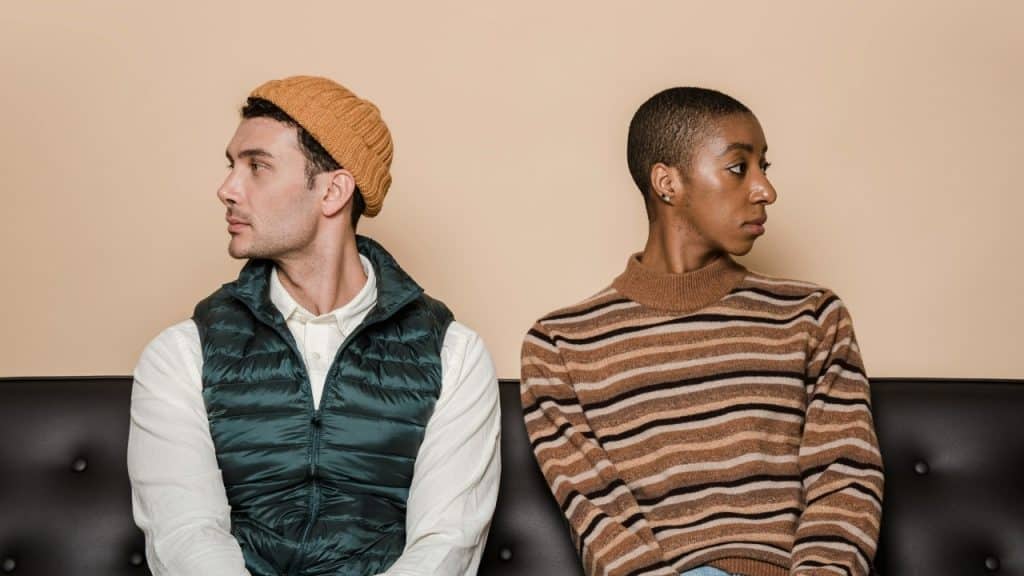 A man and woman sit on a black sofa, turned away from each other with neutral expressions.