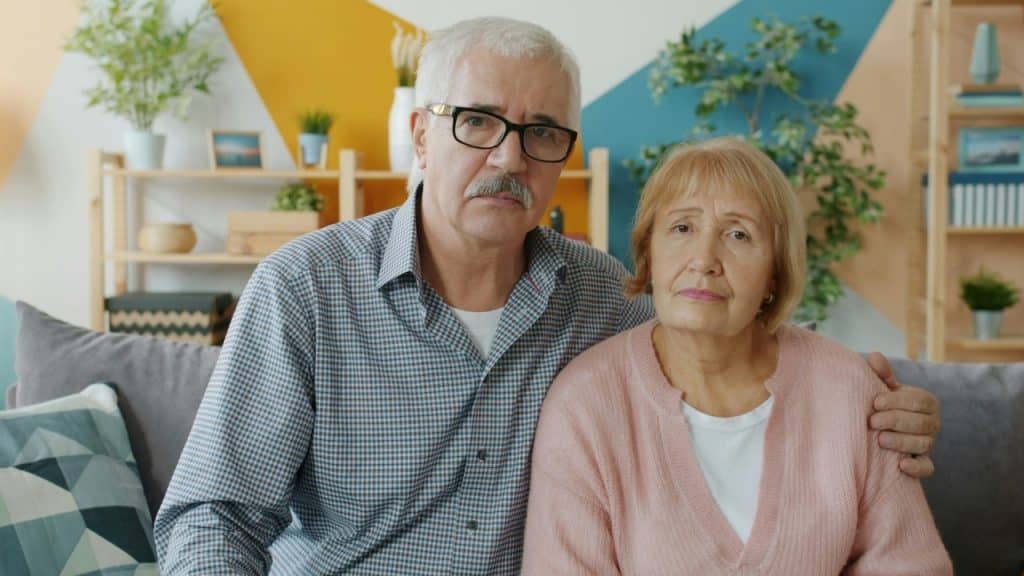 An older man and woman sit together on a sofa, looking directly at the camera with neutral expressions.