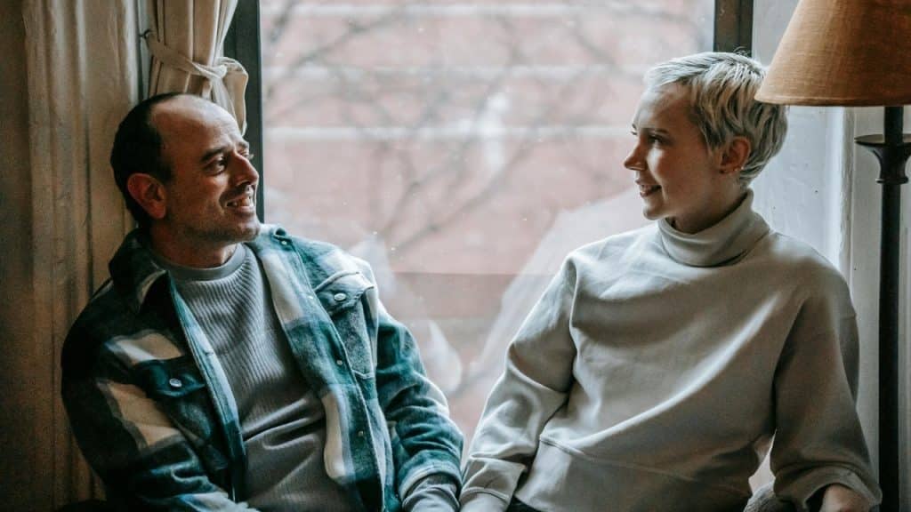 A man and woman sit by a window, looking at each other while they talk and smile.