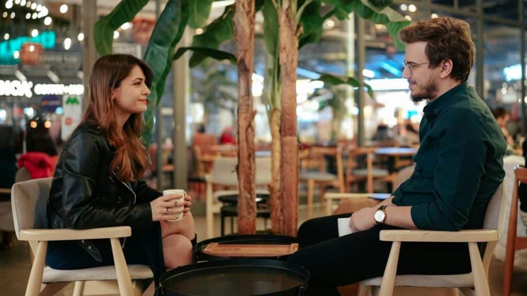 A man and woman sitting in chairs at a cafe holding mugs and looking at each other.