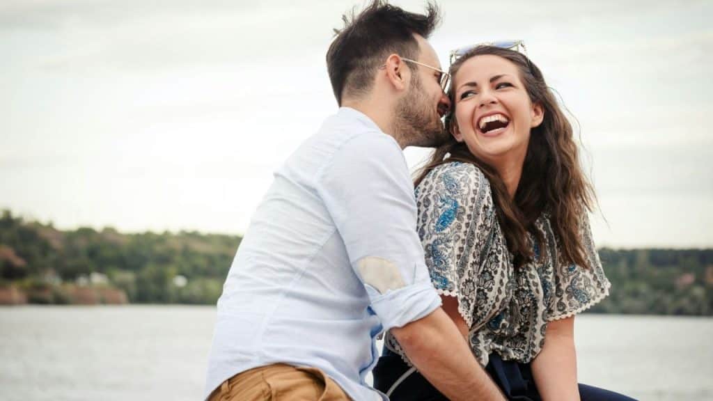 A man whispering into the ear of a laughing woman while sitting outdoors near water.