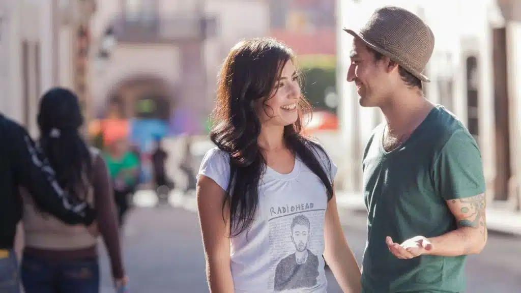 A man and woman standing on a sunny street smiling and looking at each other.