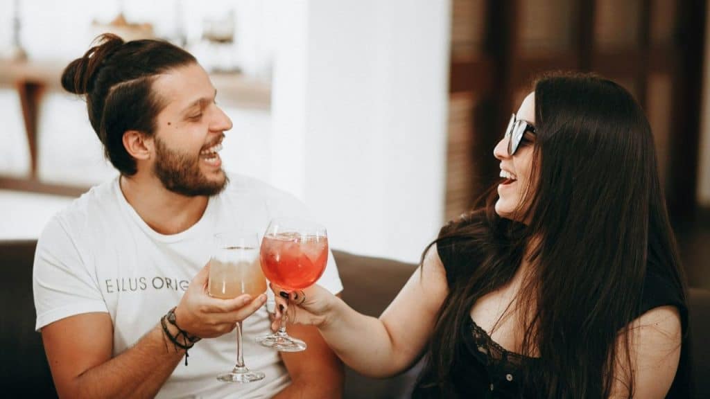 A man and woman laughing while clinking cocktail glasses together on a brown sofa.