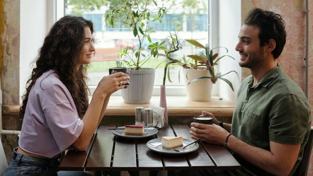 A man and woman sitting at a table by a window with coffee and cheesecake.