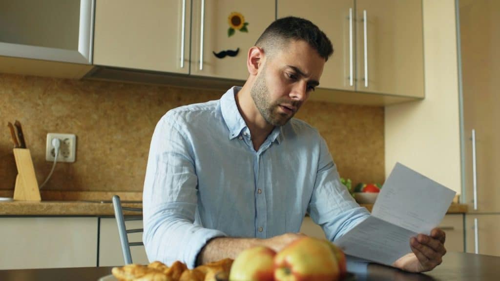 A man sits at a kitchen table while holding and looking at a white paper document.