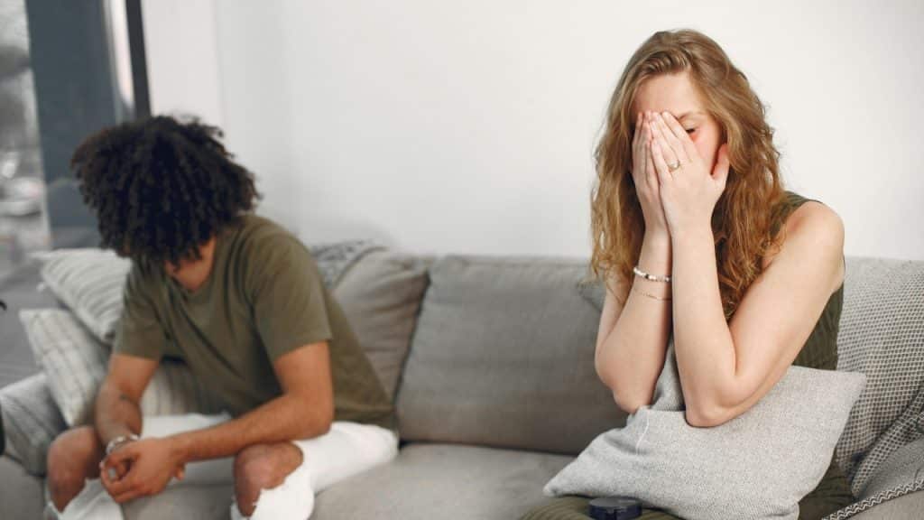 A woman covers her face with her hands while sitting on a couch near a man.