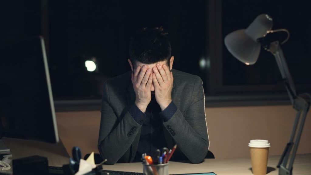 A person at a desk rests their face in their hands in a dark office setting.