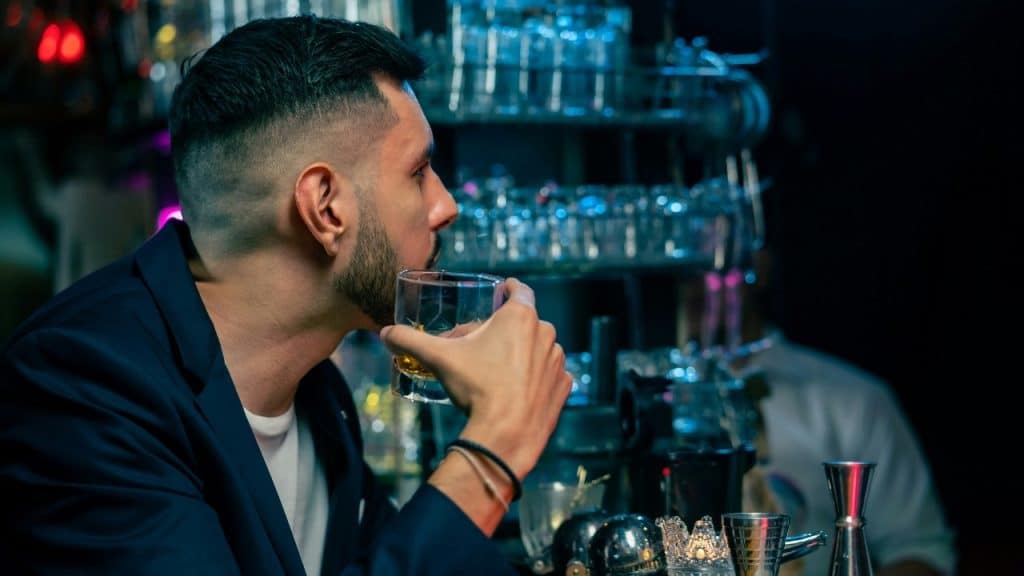 A man in a dark jacket sits at a bar holding a glass of amber liquid.