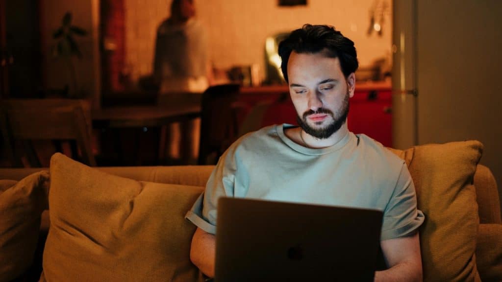 A bearded man sits on a sofa in a dimly lit room, looking at a laptop.