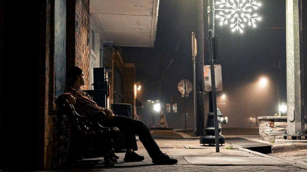 A person in a plaid shirt sits on a bench on a foggy street at night.