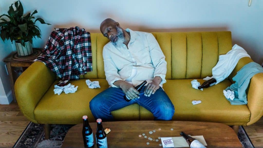 A man sleeps on a yellow sofa surrounded by glass bottles and crumpled pieces of paper.