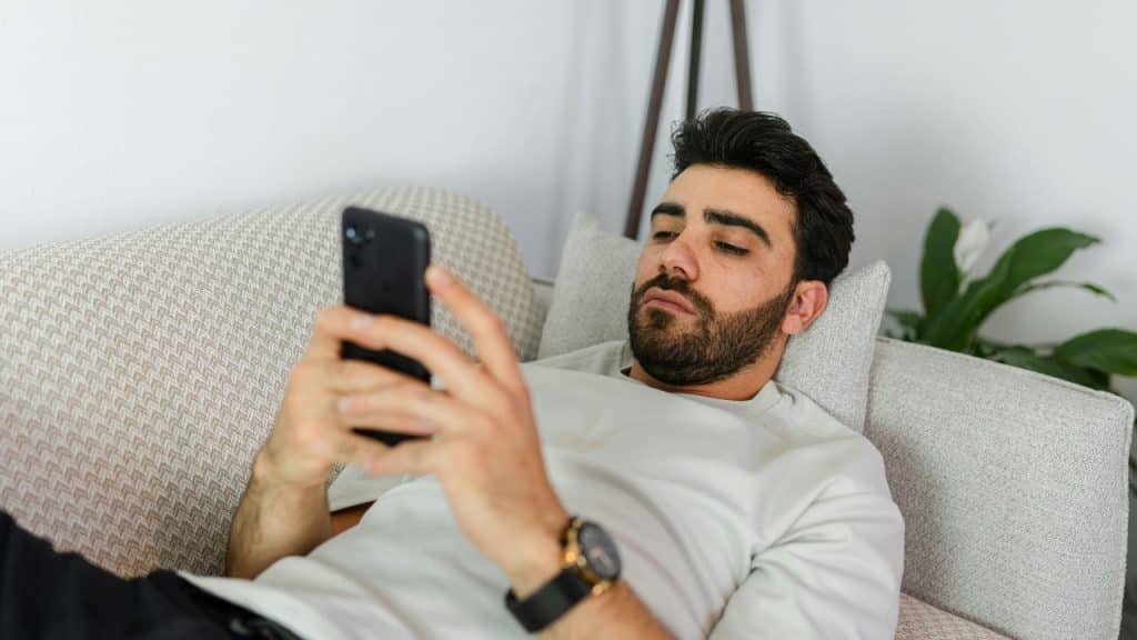 A bearded man lies on a light-colored couch while looking at a black smartphone.