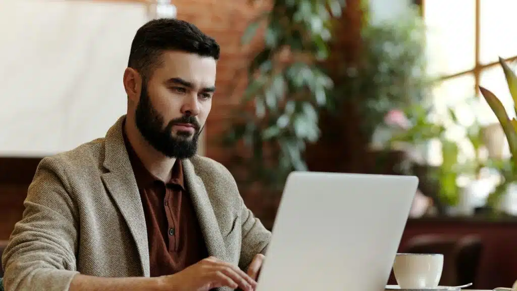 A bearded man wearing a blazer looks intently at a laptop screen in a bright room.