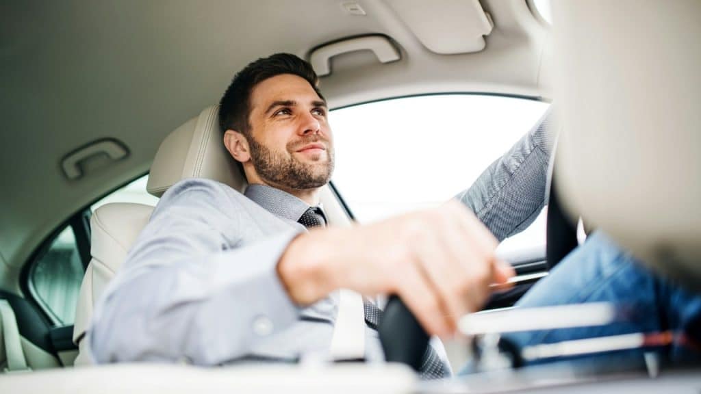 A man wearing a button-down shirt and tie smiles while sitting in a white car.