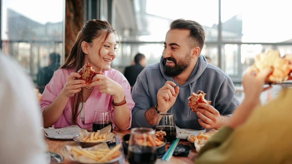 A man and woman sit at a table smiling while eating burgers and french fries.