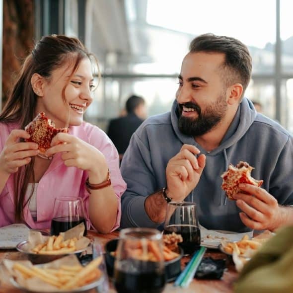 A man and woman sit at a table smiling while eating burgers and french fries.