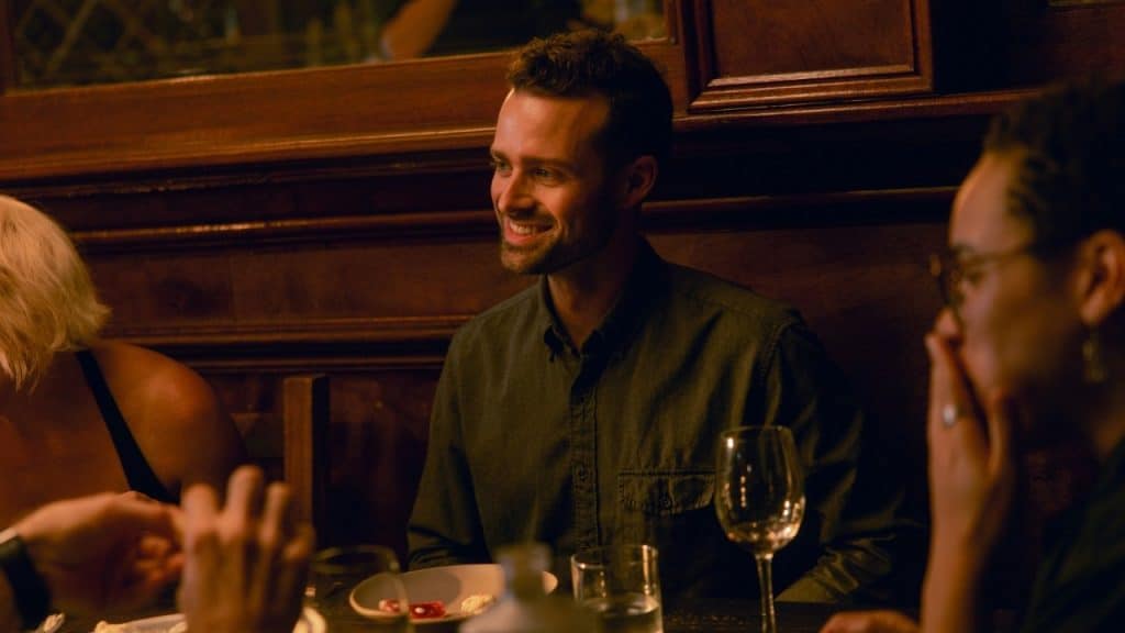 A man smiles while sitting at a dinner table with other people in dim lighting.