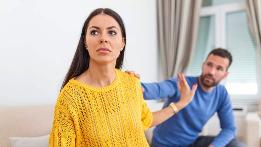 A woman in a yellow sweater looks away while a man reaches toward her shoulder.