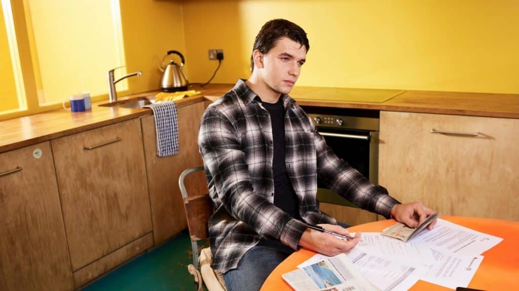 A man in a plaid shirt sits at an orange table looking at various documents.
