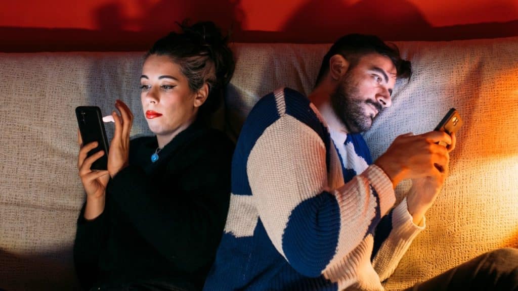 A man and woman sit back to back on a couch while using their smartphones.