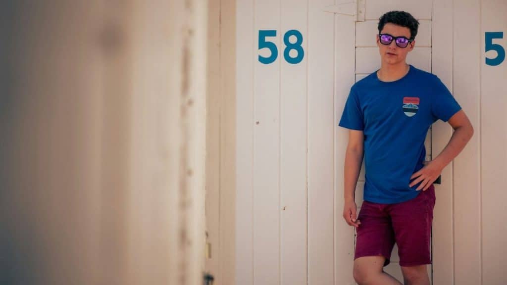 A young man wearing sunglasses and a blue shirt leans against a white wall with numbers.