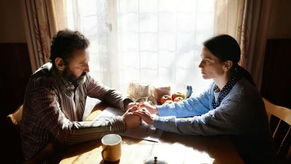 A man and woman sit at a wooden table holding hands in front of a window.
